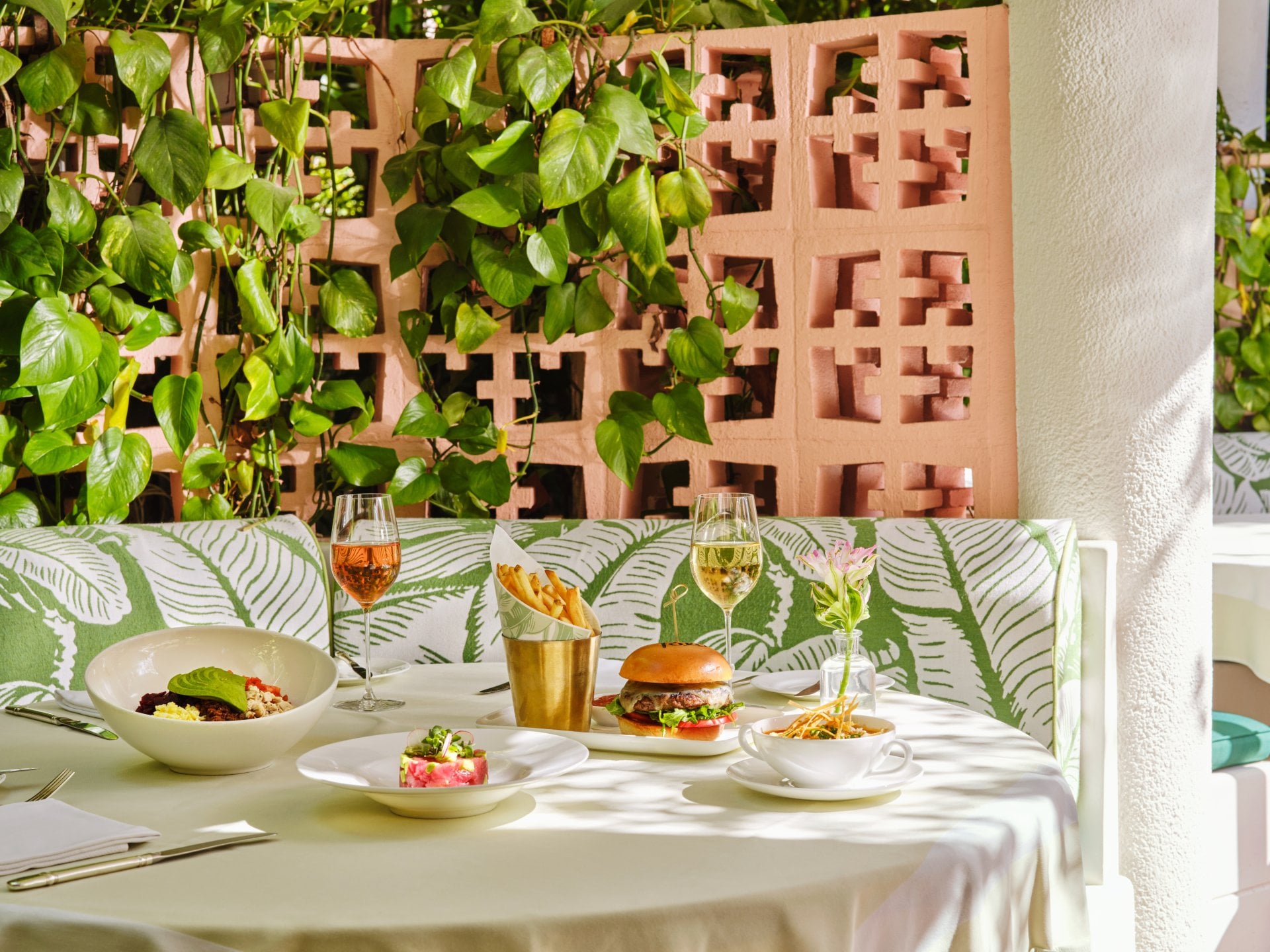 Outdoor table set with drinks, food, and leafy decor in bright, relaxed atmosphere