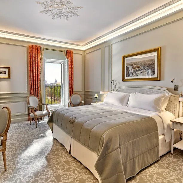 Chambre d'hôtel luxueuse avec un lit moelleux, des fauteuils élégants et une fenêtre ouverte offrant une vue panoramique. La chambre présente une décoration raffinée et des tons chauds et accueillants.