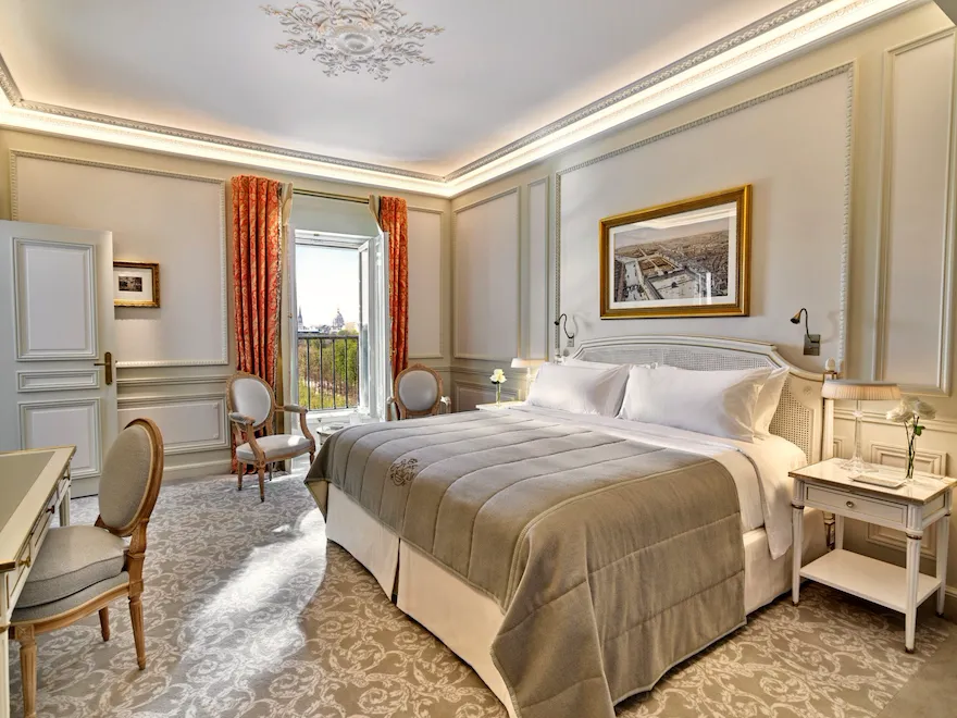 Chambre d'hôtel luxueuse avec un lit moelleux, des fauteuils élégants et une fenêtre ouverte offrant une vue panoramique. La chambre présente une décoration raffinée et des tons chauds et accueillants.