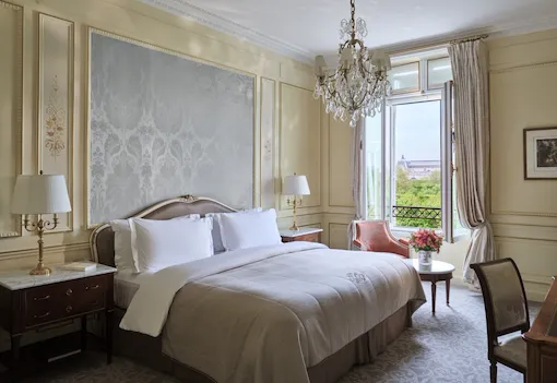 La Suite Héritage vue du côté droit du lit, à côté d’une chaise et d’une table avec des fleurs sur le dessus, devant la fenêtre, au Meurice, Paris