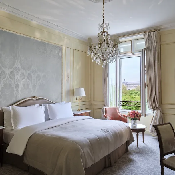 La Suite Héritage vue du côté droit du lit, à côté d’une chaise et d’une table avec des fleurs sur le dessus, devant la fenêtre, au Meurice, Paris